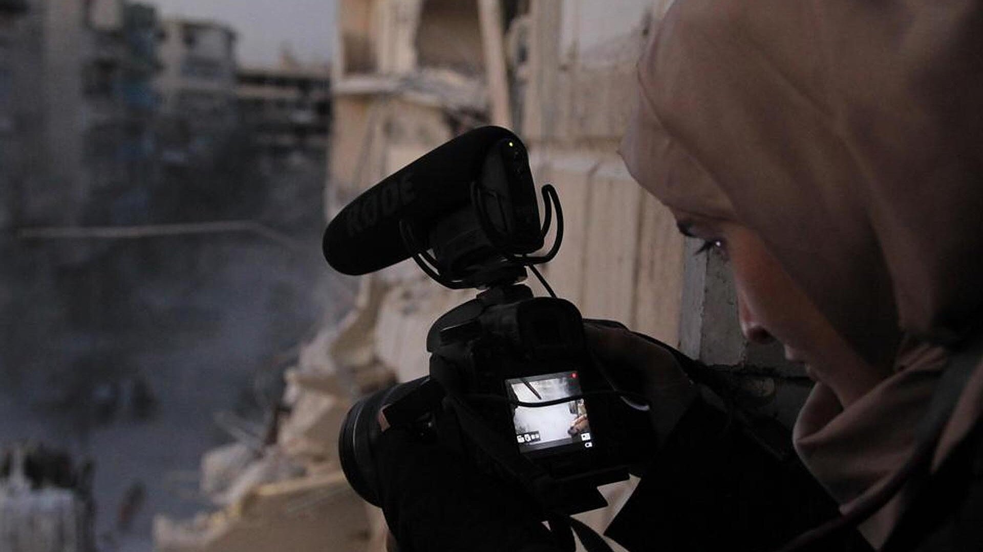 A young women holds a camera in a war torn town.